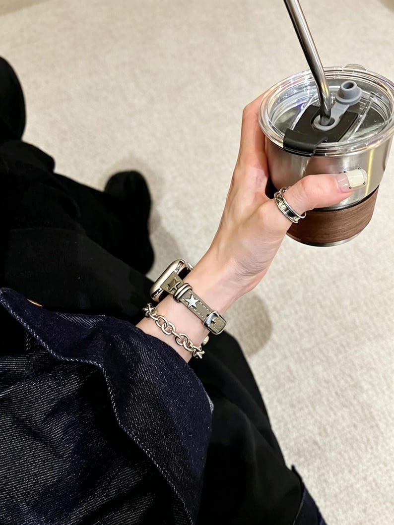 May include: A smartwatch with a grey leather strap featuring silver star studs, paired with a silver-toned chain link bracelet. The watch is worn on a wrist alongside a silver ring, while holding a stainless steel tumbler with a dark wood accent.