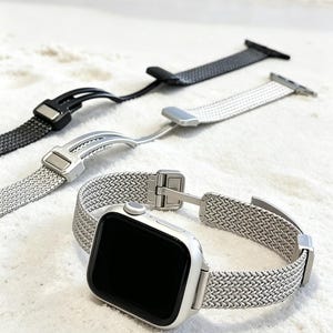 May include: A silver Apple Watch with a mesh band is displayed with two additional watch bands. One band is silver, and the other is black. The bands are on a white sandy surface with the ocean in the background.