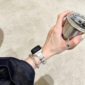 May include: A silver-toned smartwatch with a light-colored band featuring embossed star motifs, paired with a chunky silver-toned chain link bracelet and a simple silver ring, worn alongside a metallic tumbler with a clear lid and straw.