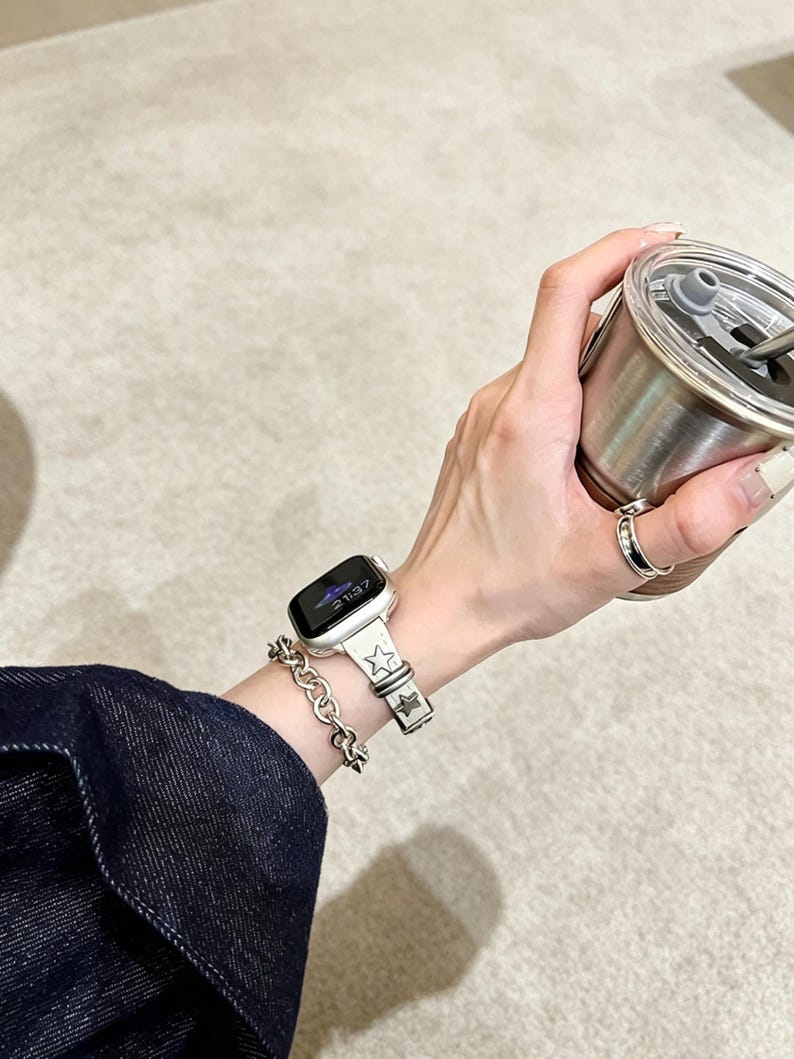 May include: A silver-toned smartwatch with a light-colored band featuring embossed star motifs, paired with a chunky silver-toned chain link bracelet and a simple silver ring, worn alongside a metallic tumbler with a clear lid and straw.