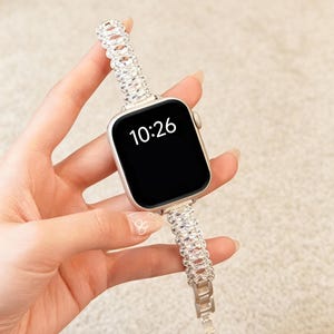 以下が含まれることがあります： 時刻10:26を表示する黒い画面のシルバーカラーのスマートウォッチ。時計には、金属製のフレームにセットされた複数の透明な楕円形のファセットカットクリスタルで構成された、チェーンのような外観の装飾的なバンドが付いています。