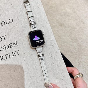 May include: A smartwatch with a rectangular screen displaying the time 21:08 and a pink orb logo. The watch features a slim, light gray leather strap adorned with metallic silver star-shaped studs and matching silver-toned metal hardware loops.