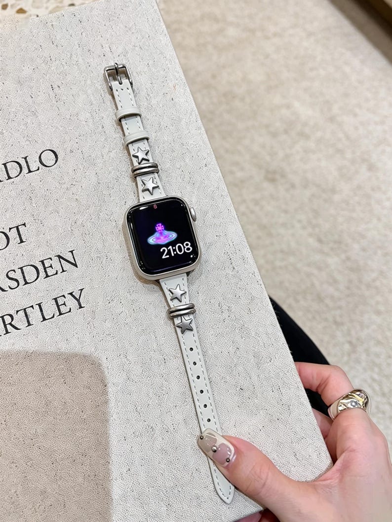 May include: A smartwatch with a rectangular screen displaying the time 21:08 and a pink orb logo. The watch features a slim, light gray leather strap adorned with metallic silver star-shaped studs and matching silver-toned metal hardware loops.