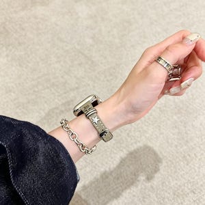 May include: A smartwatch with a light gray leather band featuring metallic star studs, paired with a silver-toned chain link bracelet. The watch band has a buckle closure and visible stitching. A silver-toned ring is worn on the finger.