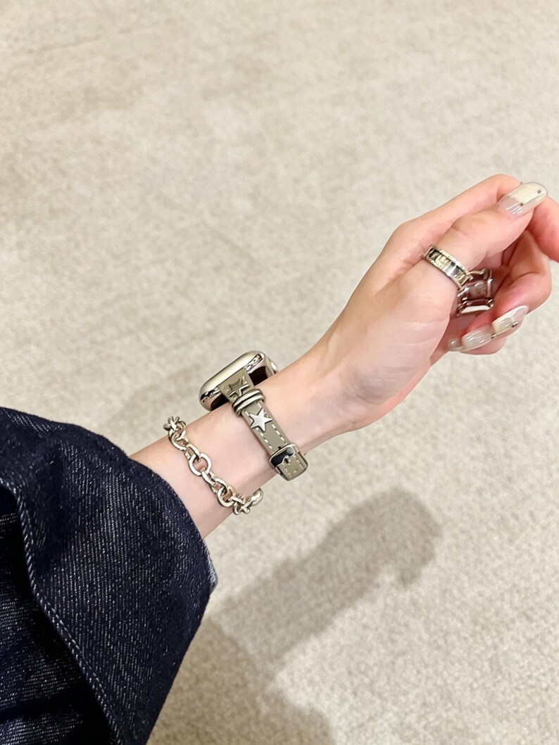 May include: A smartwatch with a light gray leather band featuring metallic star studs, paired with a silver-toned chain link bracelet. The watch band has a buckle closure and visible stitching. A silver-toned ring is worn on the finger.