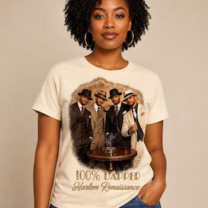 May include: A cream-colored short-sleeve t-shirt featuring a graphic print of four men in suits and fedoras standing around a table with a smoking ashtray. The text 100% DAPPER Harlem Renaissance is printed in gold lettering below the graphic design.