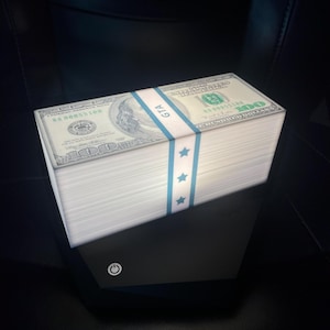 May include: A bundle of hundred-dollar bills, secured with a blue band featuring the text "GTA" and stars. The stack rests on a black electronic device with a power button symbol.