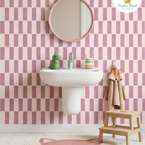 Può includere: Un bagno con carta da parati con un motivo ripetitivo di rettangoli verticali color malva e crema. Un lavabo bianco a parete si trova sotto uno specchio rotondo color malva. Un dispenser di sapone a forma di dinosauro verde e un asciugamano a forma di volpe.