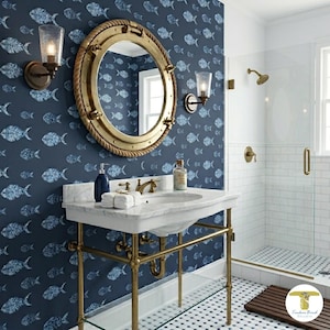May include: A bathroom vanity with a white marble countertop and brass legs, featuring a round porthole-style mirror with a rope border. The wall is covered in dark blue wallpaper with a repeating white fish pattern, complemented by two brass wall sconces.