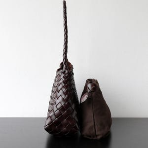 May include: Two brown leather bags. The larger bag features a woven pattern and a long, plaited handle. The smaller bag is a solid, dark brown colour with a drawstring closure. Both bags are on a black surface against a white background.
