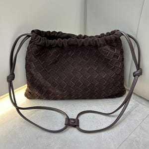 Puede incluir: Bolso de ante marr&oacute;n oscuro con un patr&oacute;n tejido. Tiene una correa larga y fina para llevar al hombro. La correa se sujeta con nudos a cada lado. El bolso se cierra con un cord&oacute;n. Dimensiones: aproximadamente 30 cm de ancho.
