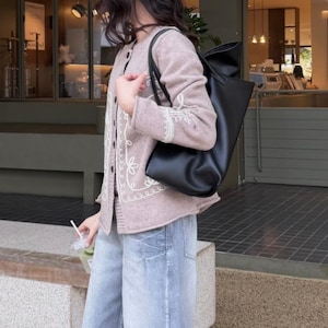 May include: A light mauve cardigan with white floral embroidery and a black tote bag. The cardigan has a button-up front and is paired with light blue denim jeans. The tote bag is made of a smooth, matte material.