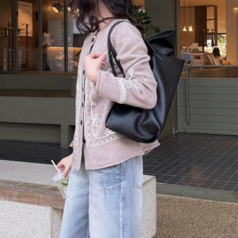 May include: A light mauve cardigan with white floral embroidery and a black tote bag. The cardigan has a button-up front and is paired with light blue denim jeans. The tote bag is made of a smooth, matte material.