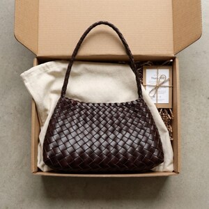 May include: A dark brown woven leather handbag with a braided handle sits in an open cardboard box. The bag is rectangular with a rounded top. A cream cloth and a small card are also in the box.
