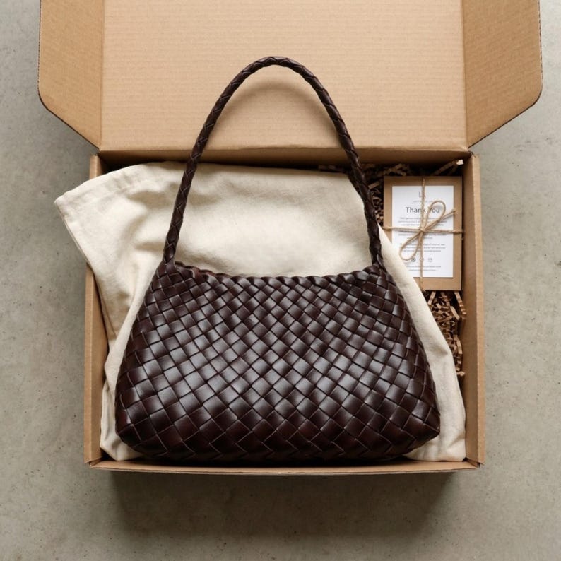May include: A dark brown woven leather handbag with a braided handle sits in an open cardboard box. The bag is rectangular with a rounded top. A cream cloth and a small card are also in the box.