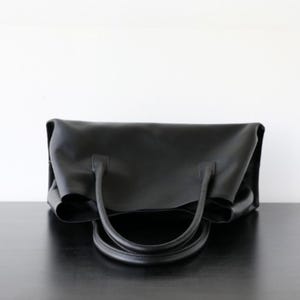 May include: A black tote bag with two curved handles. The bag is made of a smooth material and sits on a dark surface. The bag's design is simple and elegant, suitable for everyday use.