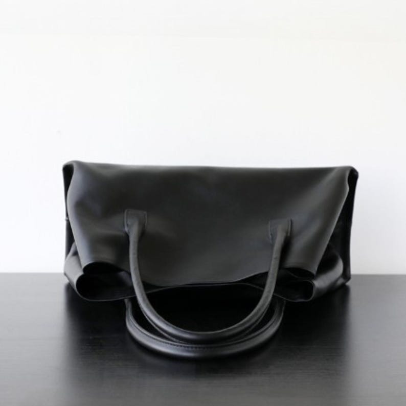 May include: A black tote bag with two curved handles. The bag is made of a smooth material and sits on a dark surface. The bag's design is simple and elegant, suitable for everyday use.
