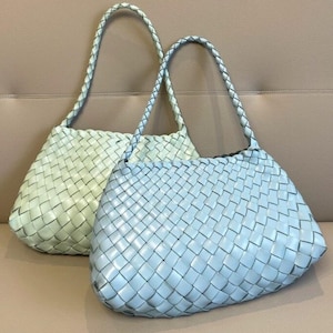 May include: Two woven leather handbags in pastel shades. One is a pale green, the other a light blue. Both have plaited handles and a basket weave pattern. The bags are displayed on a neutral-toned surface.