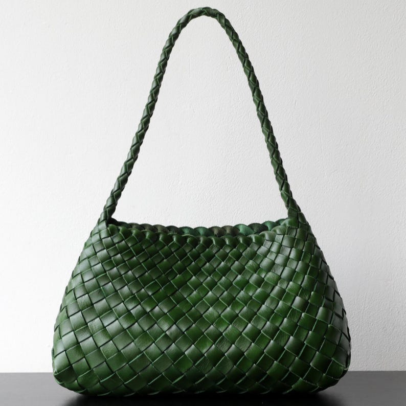 May include: A dark green woven leather handbag with a plaited handle. The bag has a rounded shape and a textured surface. The handle is long enough to be worn over the shoulder. The bag is set against a white background.