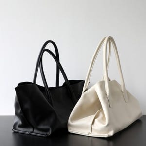 May include: Two leather tote bags. One is black, the other is off-white. Both have long, curved handles. The bags are sitting on a dark surface against a white background. The black bag is on the left, and the off-white bag is on the right.