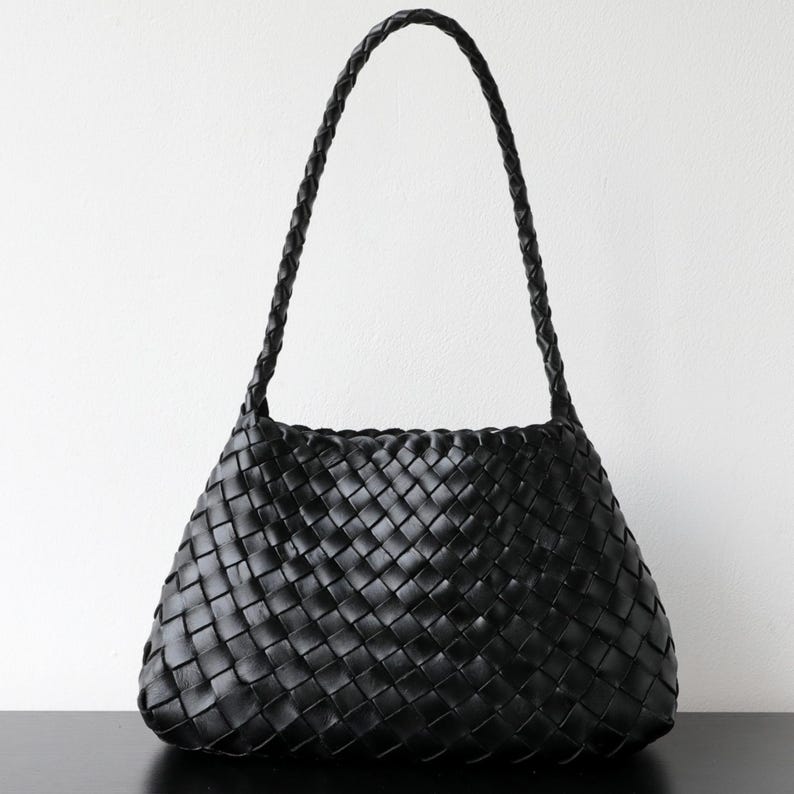 May include: Black woven leather shoulder bag with a braided handle. The bag has a trapezoid shape and a woven pattern throughout the body. The handle is also braided and appears sturdy. The bag is set against a white background.