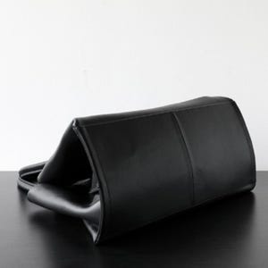 May include: A black leather tote bag with a rectangular shape. The bag has a structured design with a top handle and a smooth, glossy finish. The bag is lying on a black surface.