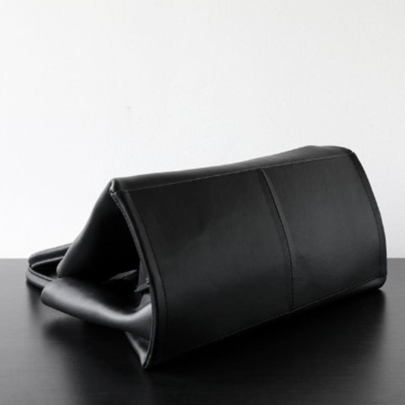 May include: A black leather tote bag with a rectangular shape. The bag has a structured design with a top handle and a smooth, glossy finish. The bag is lying on a black surface.
