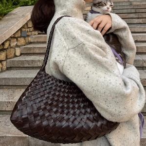 May include: A dark brown woven leather shoulder bag with a braided strap. The bag has a triangular shape and is carried over the shoulder. The bag is made of a dark brown leather material.