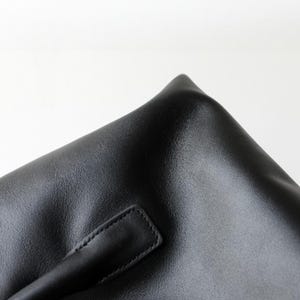 May include: Close-up of a black leather bag with a handle. The bag has a textured surface and a rectangular handle stitched to the side. The corner of the bag is visible, showing a sharp, folded edge.