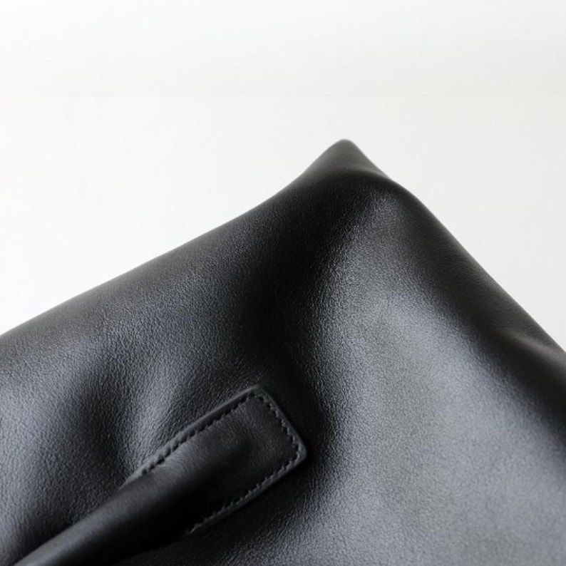 May include: Close-up of a black leather bag with a handle. The bag has a textured surface and a rectangular handle stitched to the side. The corner of the bag is visible, showing a sharp, folded edge.