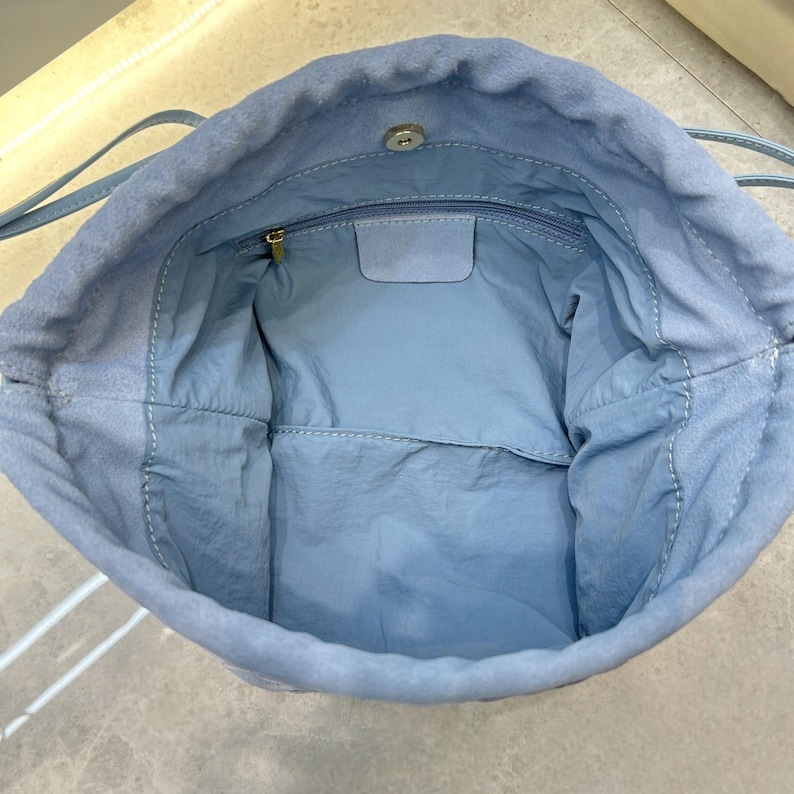 Puede incluir: Un bolso azul claro con cord&oacute;n y un exterior suave similar al ante. El interior presenta un forro de tela azul m&aacute;s claro con un bolsillo con cremallera y un cierre de bot&oacute;n magn&eacute;tico. El bolso tiene finas correas para el hombro.