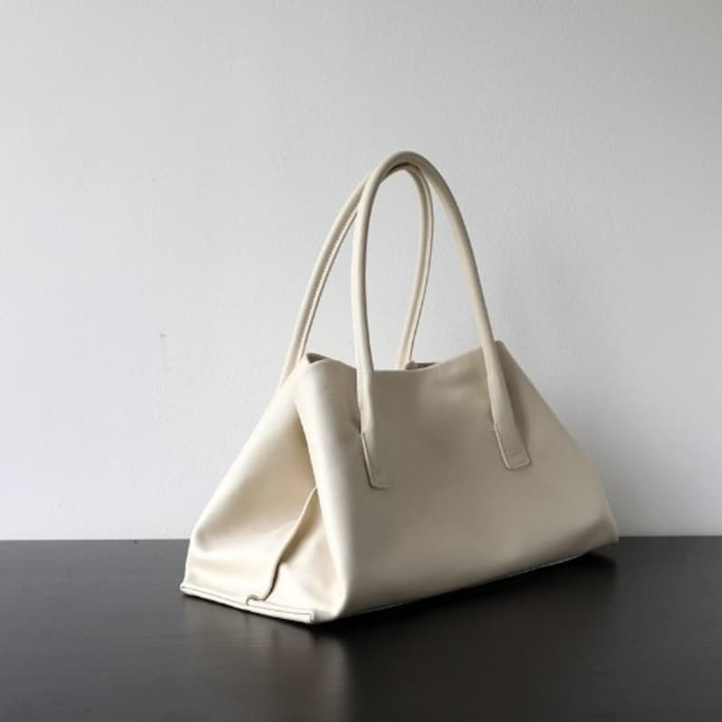 May include: Cream-colored tote bag with two long handles. The bag has a rectangular shape with a trapezoidal base. The bag is made of a smooth material and sits on a dark surface against a white wall.