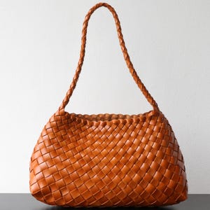 May include: A tan-brown woven leather tote bag with a braided handle. The bag has a basket-weave pattern and a rounded shape. The bag is a medium size, suitable for everyday use.