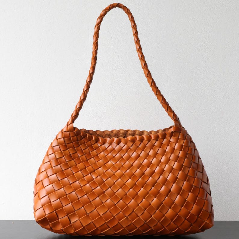 May include: A tan-brown woven leather tote bag with a braided handle. The bag has a basket-weave pattern and a rounded shape. The bag is a medium size, suitable for everyday use.
