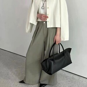 May include: A cream-colored jacket with button closures, a white top, and olive-green wide-leg pants. A black tote bag with handles is held in the hand. The outfit is paired with black pointed-toe shoes. The overall style is minimalist and modern.