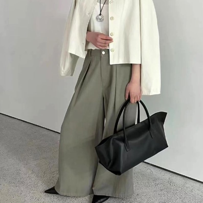 May include: A cream-colored jacket with button closures, a white top, and olive-green wide-leg pants. A black tote bag with handles is held in the hand. The outfit is paired with black pointed-toe shoes. The overall style is minimalist and modern.