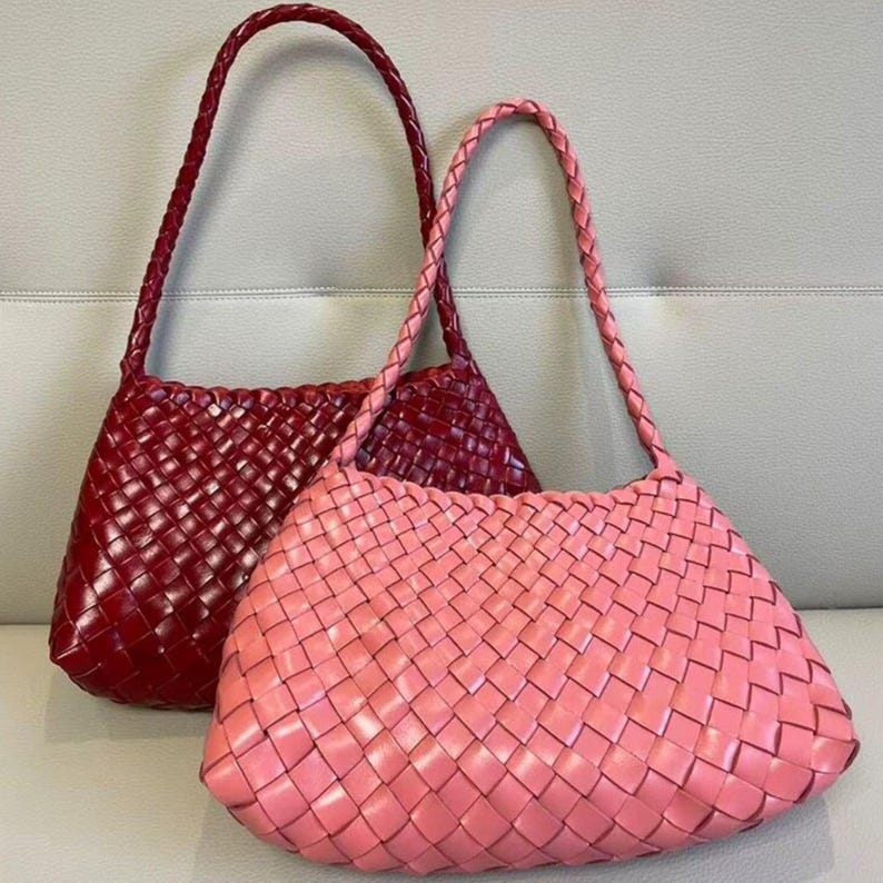 May include: Two woven leather handbags. One is a deep red, the other is a soft pink. Both bags have braided handles and a rectangular shape with a rounded top. The weave pattern is consistent throughout the bags.
