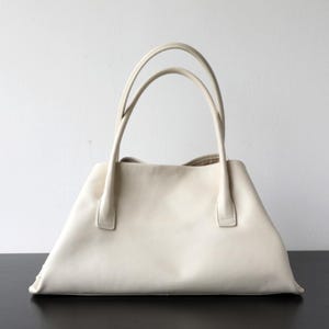 May include: Cream-colored leather tote bag with two curved handles. The bag has a trapezoidal shape with a flat base and a wide opening. The handles are attached to the top of the bag with small, rectangular pieces of leather.