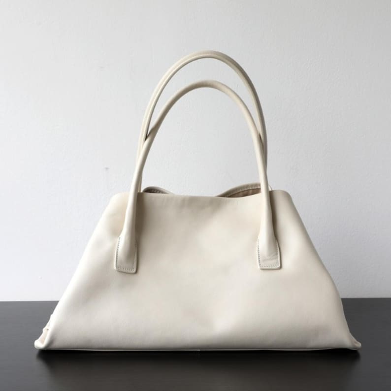 May include: Cream-colored leather tote bag with two curved handles. The bag has a trapezoidal shape with a flat base and a wide opening. The handles are attached to the top of the bag with small, rectangular pieces of leather.