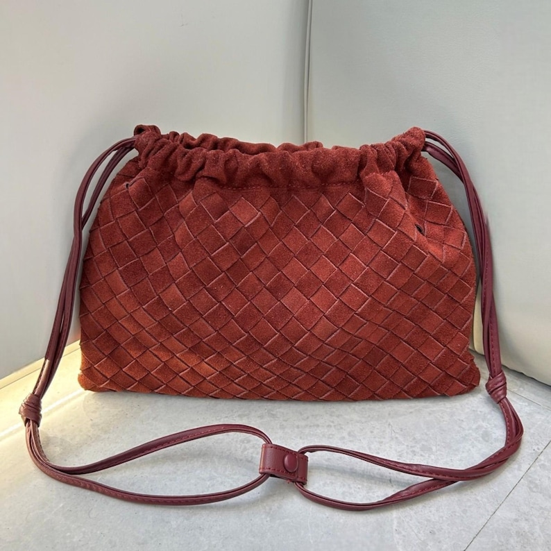 Puede incluir: Un bolso bandolera de ante rojo con dise&ntilde;o tejido. El bolso tiene un cierre de cord&oacute;n y una correa larga y ajustable. La correa est&aacute; unida al bolso con un nudo a cada lado. El bolso est&aacute; hecho de un material suave y texturizado.