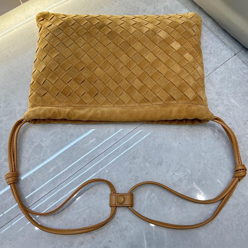 Puede incluir: Un bolso de mano de ante marr&oacute;n con un dise&ntilde;o tejido en el cuerpo principal. El bolso tiene un cierre de cord&oacute;n y correas largas y finas que se conectan con un peque&ntilde;o cierre rectangular.
