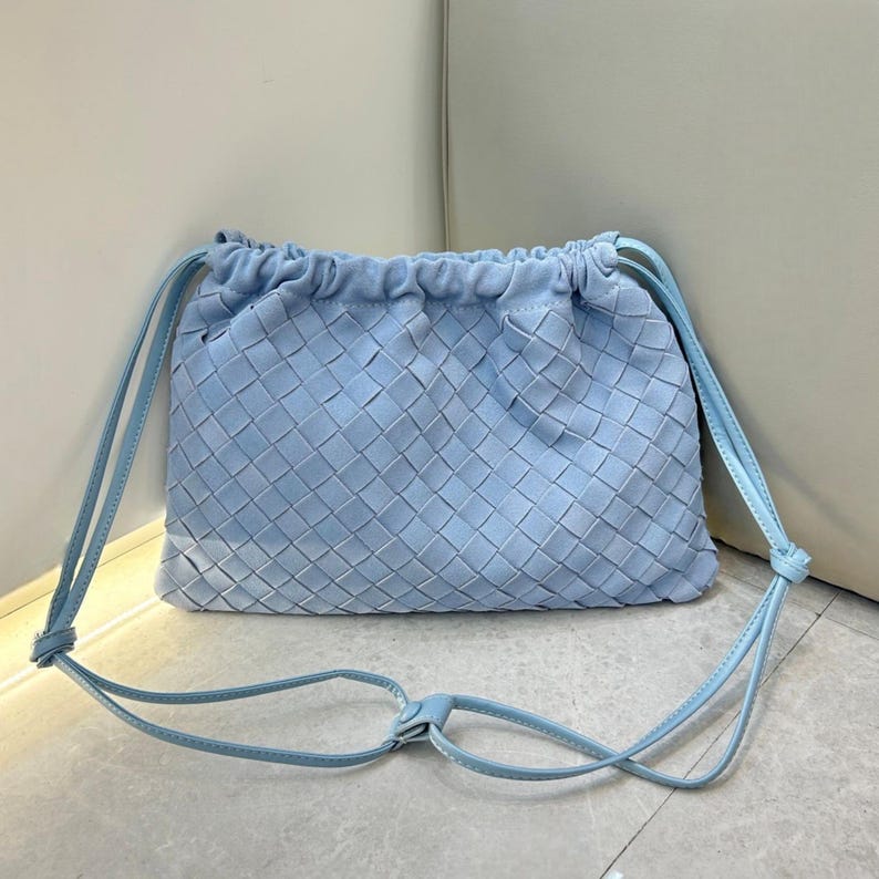 Puede incluir: Bolso bandolera de cuero tejido azul claro con cierre de cord&oacute;n. El bolso tiene forma rectangular con un patr&oacute;n tejido y una correa larga y ajustable. La correa y el cord&oacute;n son del mismo color azul claro.