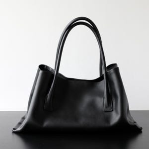 May include: A black tote bag with two long handles. The bag is made of a smooth material and has a simple, elegant design. The handles are curved and attached to the top of the bag. The bag is sitting on a black surface.