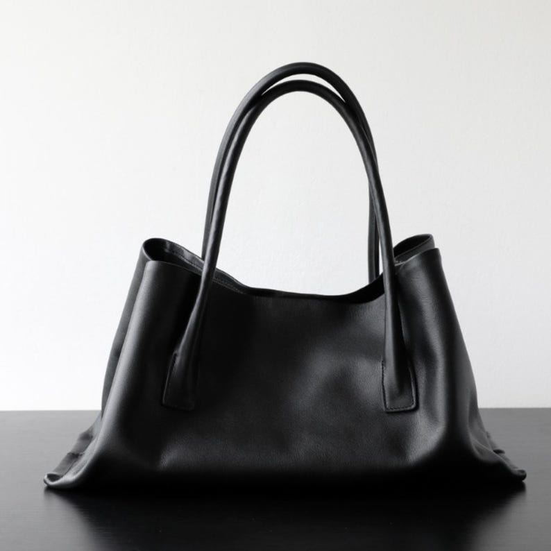 May include: A black tote bag with two long handles. The bag is made of a smooth material and has a simple, elegant design. The handles are curved and attached to the top of the bag. The bag is sitting on a black surface.