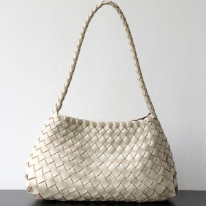 May include: Cream-coloured woven leather shoulder bag with a braided handle. The bag has a rectangular shape with a rounded base and a woven pattern. The handle is also braided and appears sturdy. The bag is made of leather.
