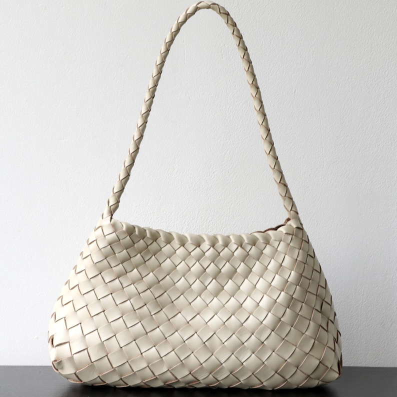May include: Cream-coloured woven leather shoulder bag with a braided handle. The bag has a rectangular shape with a rounded base and a woven pattern. The handle is also braided and appears sturdy. The bag is made of leather.
