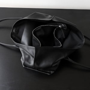 May include: Black leather tote bag with long handles, open to reveal a spacious interior and a zippered pocket. The bag is made of a soft, supple material and is designed for everyday use.