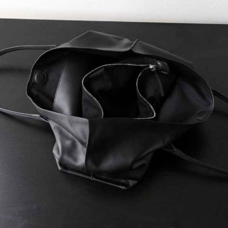 May include: Black leather tote bag with long handles, open to reveal a spacious interior and a zippered pocket. The bag is made of a soft, supple material and is designed for everyday use.