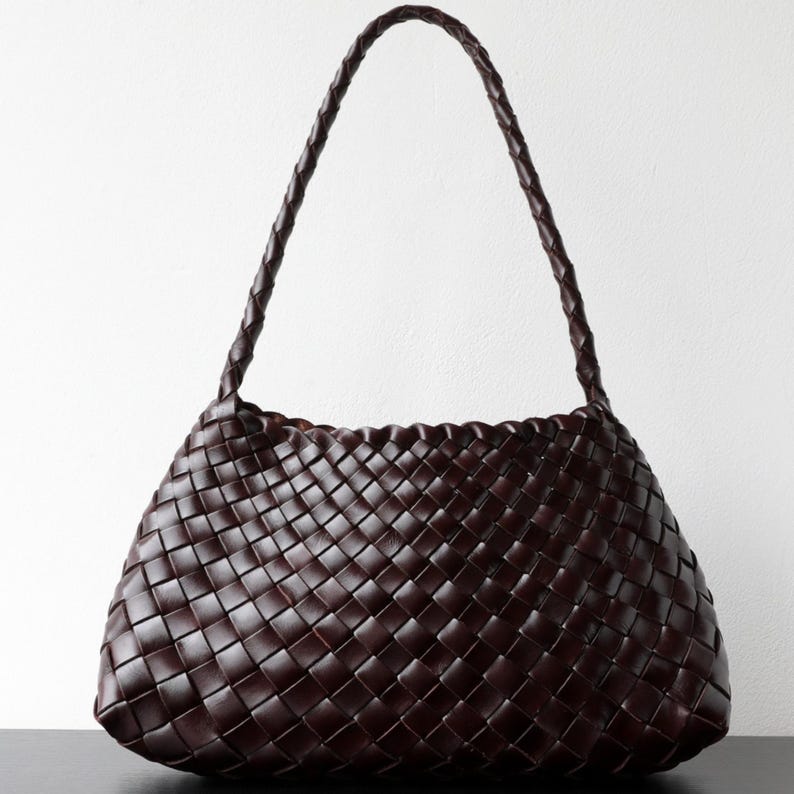 May include: A dark brown woven leather shoulder bag with a braided handle. The bag has a trapezoid shape with a woven pattern throughout the body. The handle is a single, long braid.