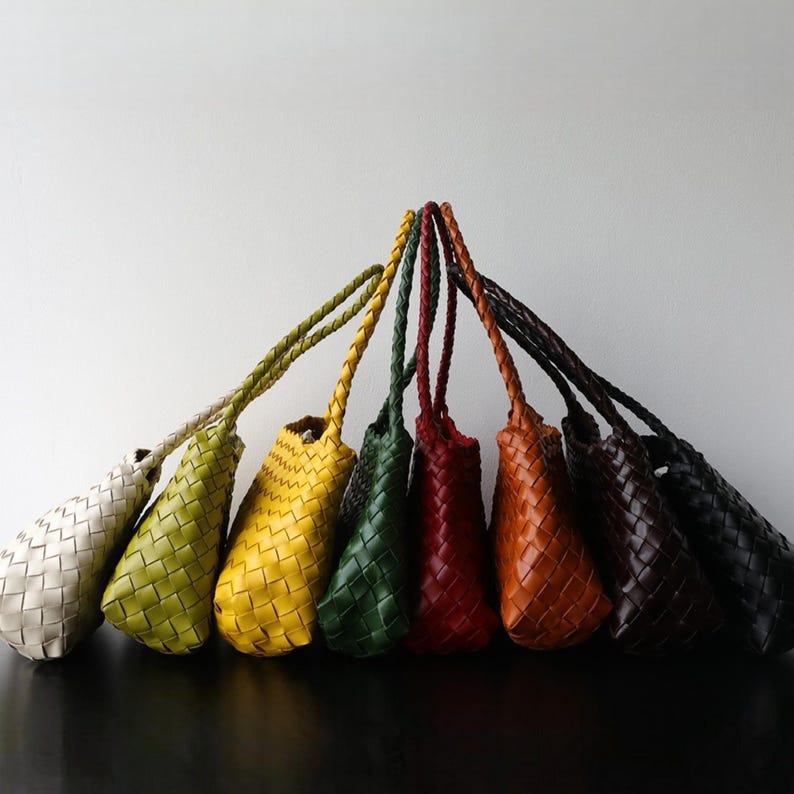 May include: A selection of woven leather tote bags in a range of colours: cream, green, yellow, dark green, red, orange, brown, and black. Each bag has a braided handle and a distinctive woven design.